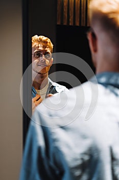 Young man in glasses looks at himself in the mirror and straightens his shirt