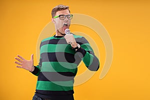 Young man in glasses singing song in microphone. Young posing isolated on yellow orange background. People lifestyle concept