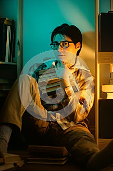 An young man with glasses sits on the floor between bookshelves in the night, holding a stack of books and looking directly at