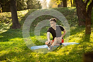 Young man in glasses training yoga outdoors. Sporty guy makes relaxing exercise on a blue yoga mat, in park. Copy space