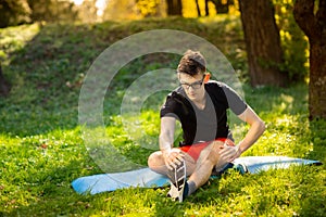 Young man in glasses training yoga outdoors. Sporty guy makes relaxing exercise on a blue yoga mat, in park. Copy space