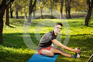 Young man in glasses training yoga outdoors. Sporty guy makes relaxing exercise on a blue yoga mat, in park. Copy space