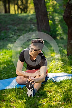 Young man in glasses training yoga outdoors. Sporty guy makes relaxing exercise on a blue yoga mat, in park. Copy space