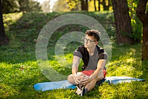 Young man in glasses training yoga outdoors. Sporty guy makes relaxing exercise on a blue yoga mat, in park. Copy space