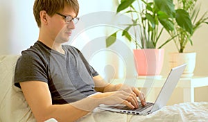 A young man with glasses works in the bedroom with a laptop.