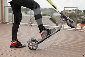 Young man in gray jeans in sneakers posing with an electric scooter on a summer terrace. Closeup of male legs on a modern scooter