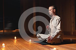 A young man in gray practice clothes sits in a lotus position with a red rosary in his hand in meditation. Dark room