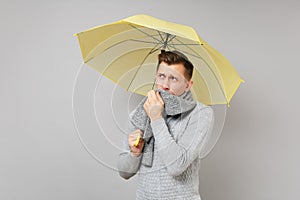 Young man in gray sweater covering mouth with scarf, looking aside, holding yellow umbrella on grey wall