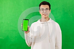 A young man with a green screen phone, wearing glasses, smiling and looking at the camera on a green background