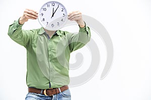 Young man in green is waiting, holding clock in front of his face over white background.