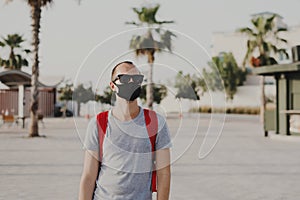 Young man in grey t-shirt, sun glasses and with backpack wearing black protective mask