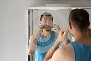 Young man grimaces and makes faces to his reflection in mirror in bathroom. View over the shoulder