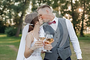 Young man groom kissing young attractive woman bride, stretching hands with glasses of wine. Wedding, celebration.