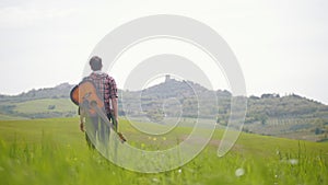 A young man guitarist walking on a bright green meadow - traveling concept