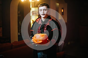 Young man in a Halloween costume of Count Dracula raises a carved pumpkin
