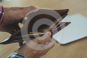 Young man hands holding brown opened empty wallet on wooden table with laptop background. He was in financial trouble after being
