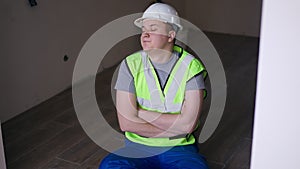Young man in hard hat and green vest sitting on floor in new apartment thinking. Portrait of smiling positive Caucasian