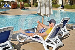 Young man in hat and black sunglasses lying on a sun lounger at the hotel near sweeming pool. Relax in summer pool