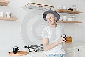 Young man in hat smiles drink fresh coffee in ceramic black mug. Bright kitchen background