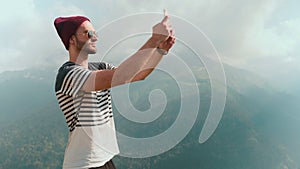 A young man in a hat and sunglasses, standing against the background of mountains and photographing beautiful landscapes