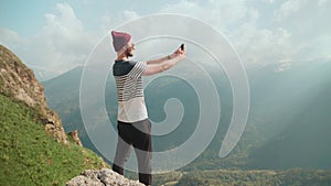 A young man in a hat and sunglasses, standing against the background of mountains and photographing beautiful landscapes