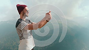 A young man in a hat and sunglasses, standing against the background of mountains and photographing beautiful landscapes