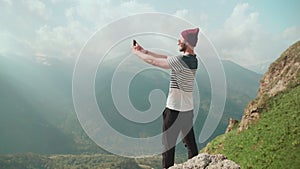 A young man in a hat and sunglasses, standing against the background of mountains and photographing beautiful landscapes