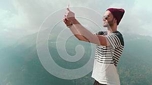 A young man in a hat and sunglasses, standing against the background of mountains and photographing beautiful landscapes