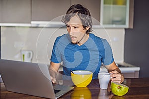 Young man having breakfast and using laptop on the kitchen