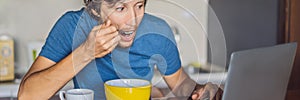 Young man having breakfast and using laptop on the kitchen BANNER, LONG FORMAT