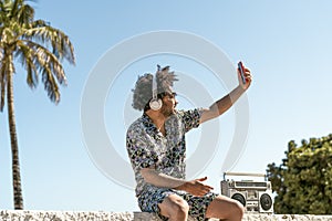 Young man having fun taking selfie with mobile smartphone while listening music with headphones and vintage boombox during summer