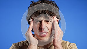 Young man having headache, studio portrait. Guy puts hand on head on blue