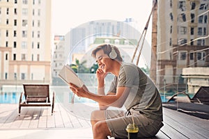 Young man with headphone near swimming pool reading book