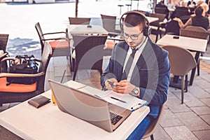 Young man with headphones and glasses using smart phone and laptop computer in street cafe