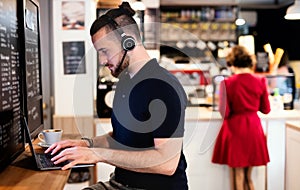 Young man with headphones and laptop indoors in cafe, working.