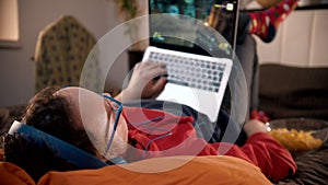 Young man in headphones lying on the bed playing shooter game on the laptop