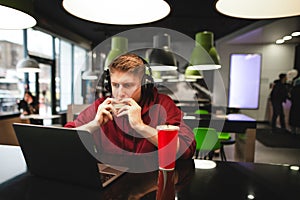 Young man in the headphones sits at the table in the cafe, eats a tasty burger and looks at the laptop screen