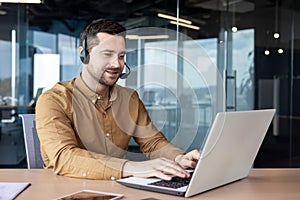 A young man in a headset sits in the office and works on a laptop. types on the keyboard, chats, talks on a video call