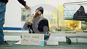 Young man helps to homeless person and giving him some food while beggar drink alcohol and sit near shopping cart at