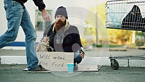 Young man helps to homeless person and giving him some money while beggar drink alcohol and sit near shopping cart at