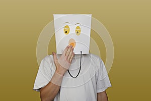 Young man hiding face behind paper bag with drawn emoticon on light background