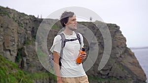 A young man on a hike among rocks near the sea. High wind conditions, big waves on the sea. He drinks from an orange