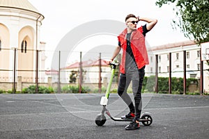 Young man hipster in sneakers in a stylish red denim vest in black jeans in sunglasses stands with an electric scooter