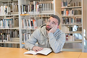 Joven hombre su cabeza está descansando sobre su mano cansado aburrido de lectura un libro en biblioteca 