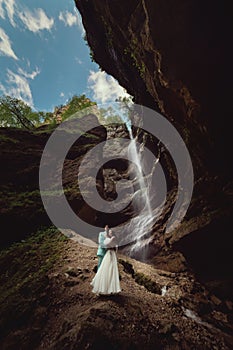 A young man and his wife are standing in an embrace in a canyon against the backdrop of a high waterfall. Newlyweds