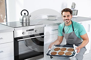 Young man holding baking sheet with cookies near oven