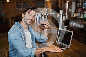 Young man holding beer while sitting by laptop in pub