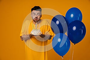 Young man holding birthday cake and blowing out candles while standing with blue air balloons isolated