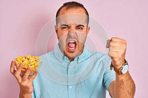 Young man holding bowl with macaroni pasta standing over isolated pink background annoyed and frustrated shouting with anger,