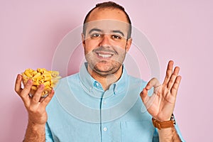 Young man holding bowl with macaroni pasta standing over isolated pink background doing ok sign with fingers, excellent symbol